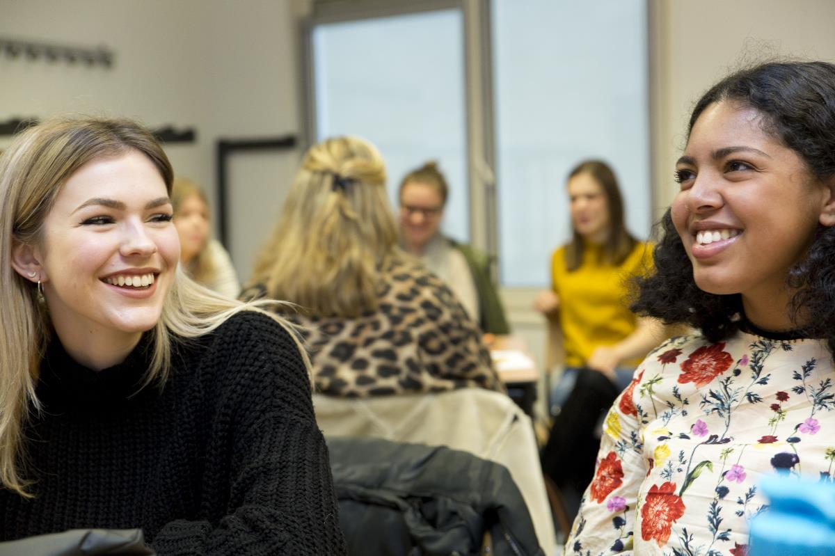 Twee vrouwelijke studenten lachen met op de achtergrond een groep studenten aan het werk