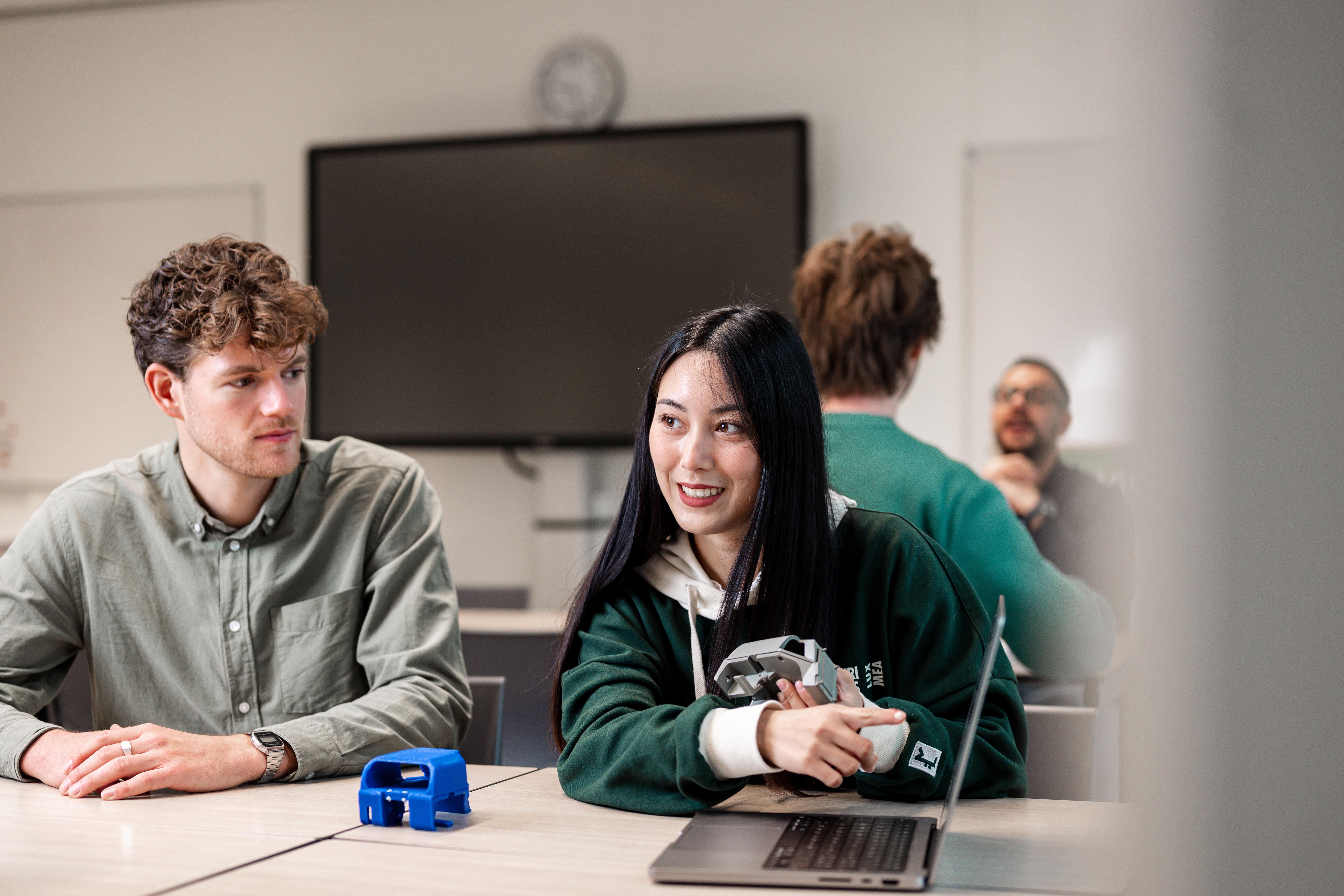 Twee studenten overleggen samen aan een tafel in een klaslokaal, met technische onderdelen en een laptop voor zich.