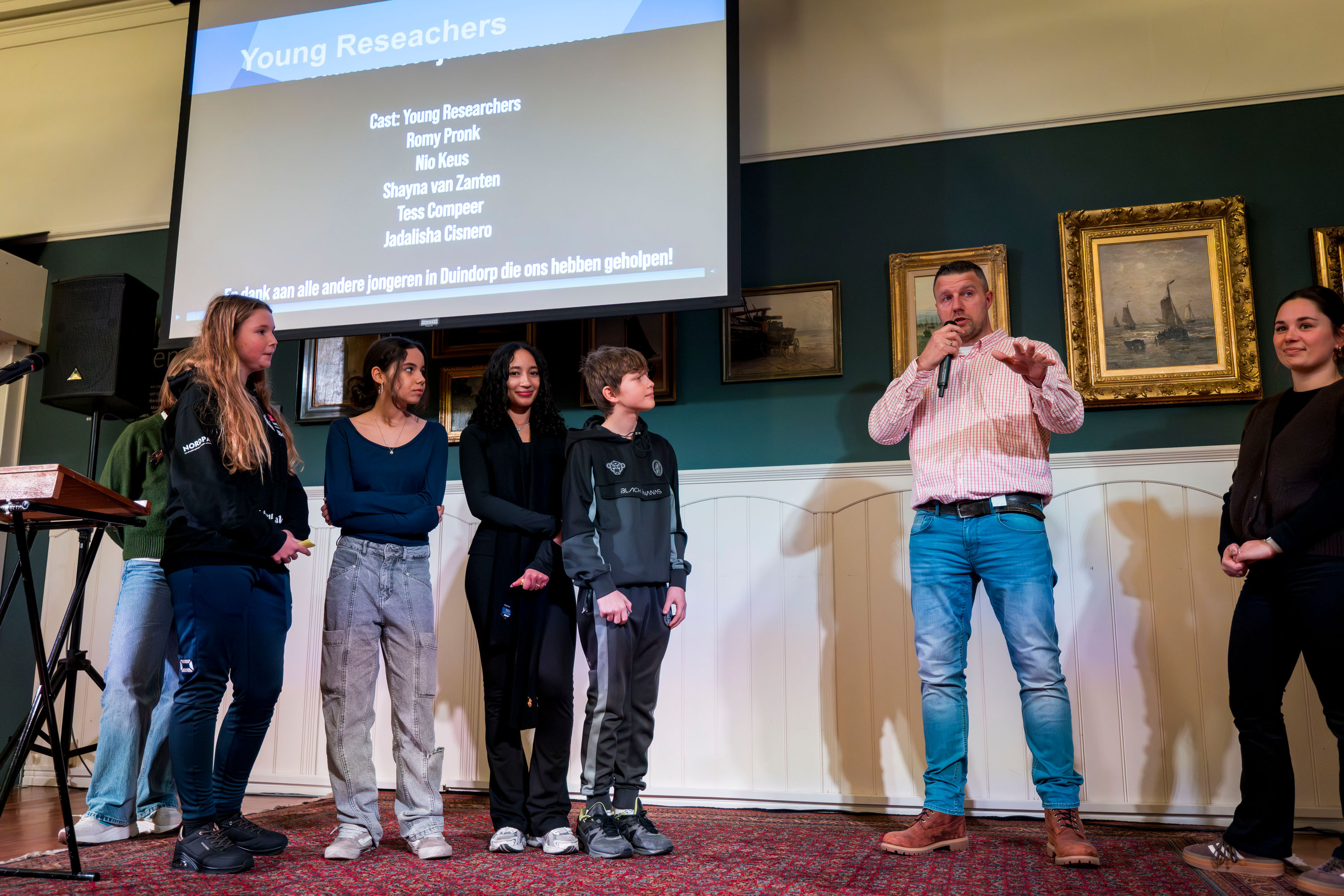 A group of young people stand on a stage while an adult speaks into a microphone, with a screen behind them displaying “Young Researchers” and a list of names.