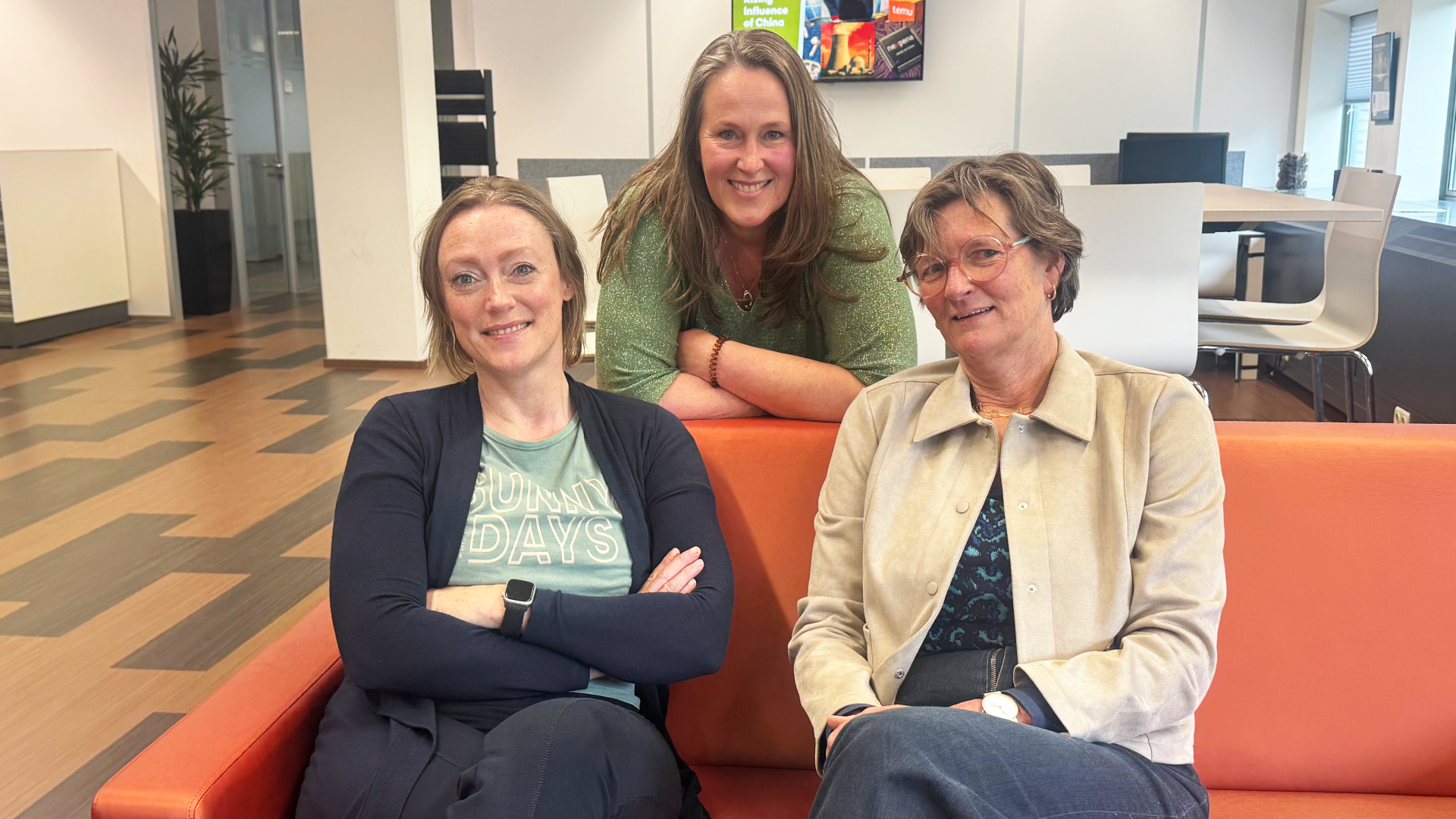 Three people seated and standing around an orange couch in a bright office space.