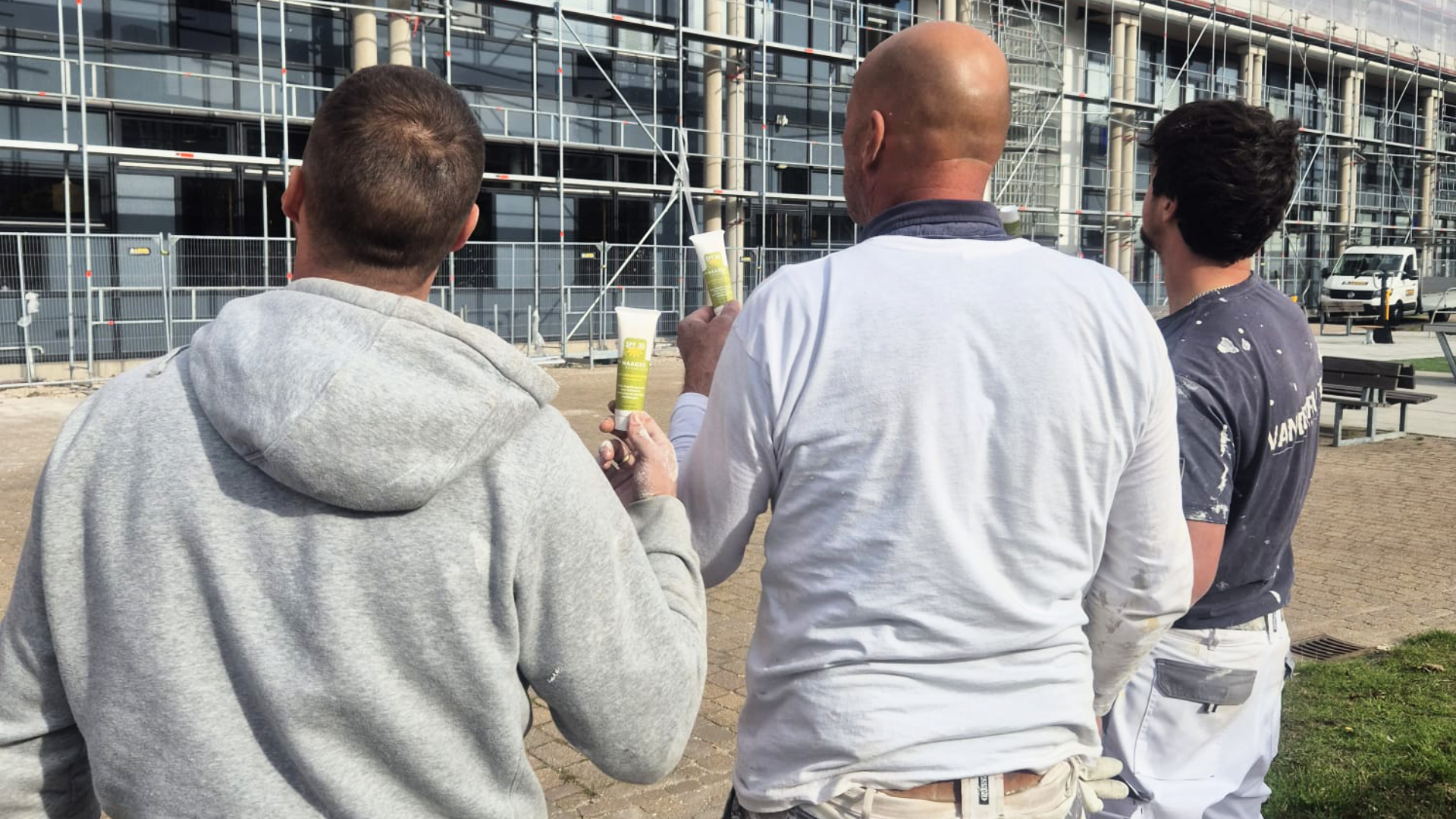 Three construction workers look at a building under construction with scaffolding, holding tubes of material.