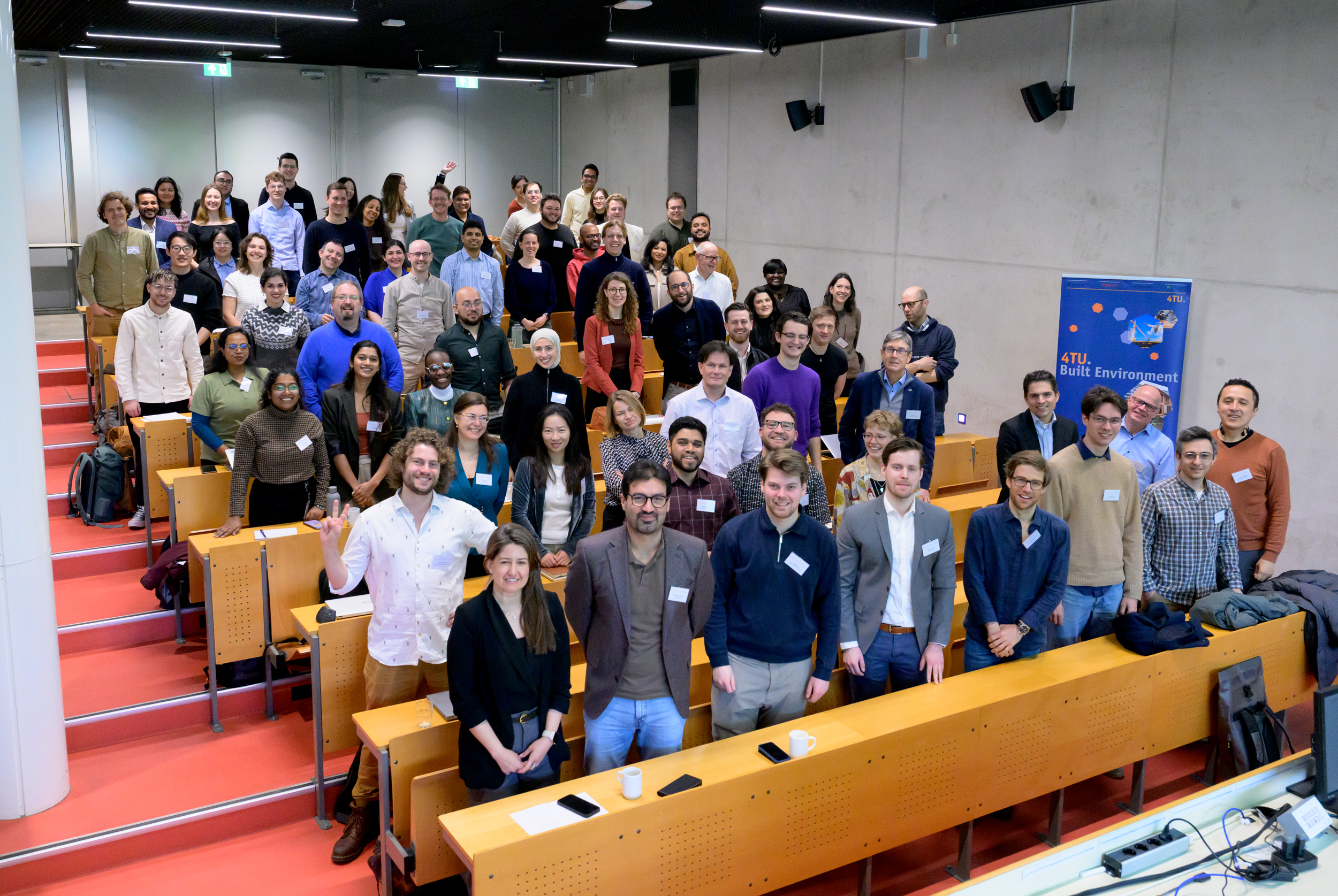 Grote groep mensen poseert in een collegezaal met een 4TU‑banner op de achtergrond.