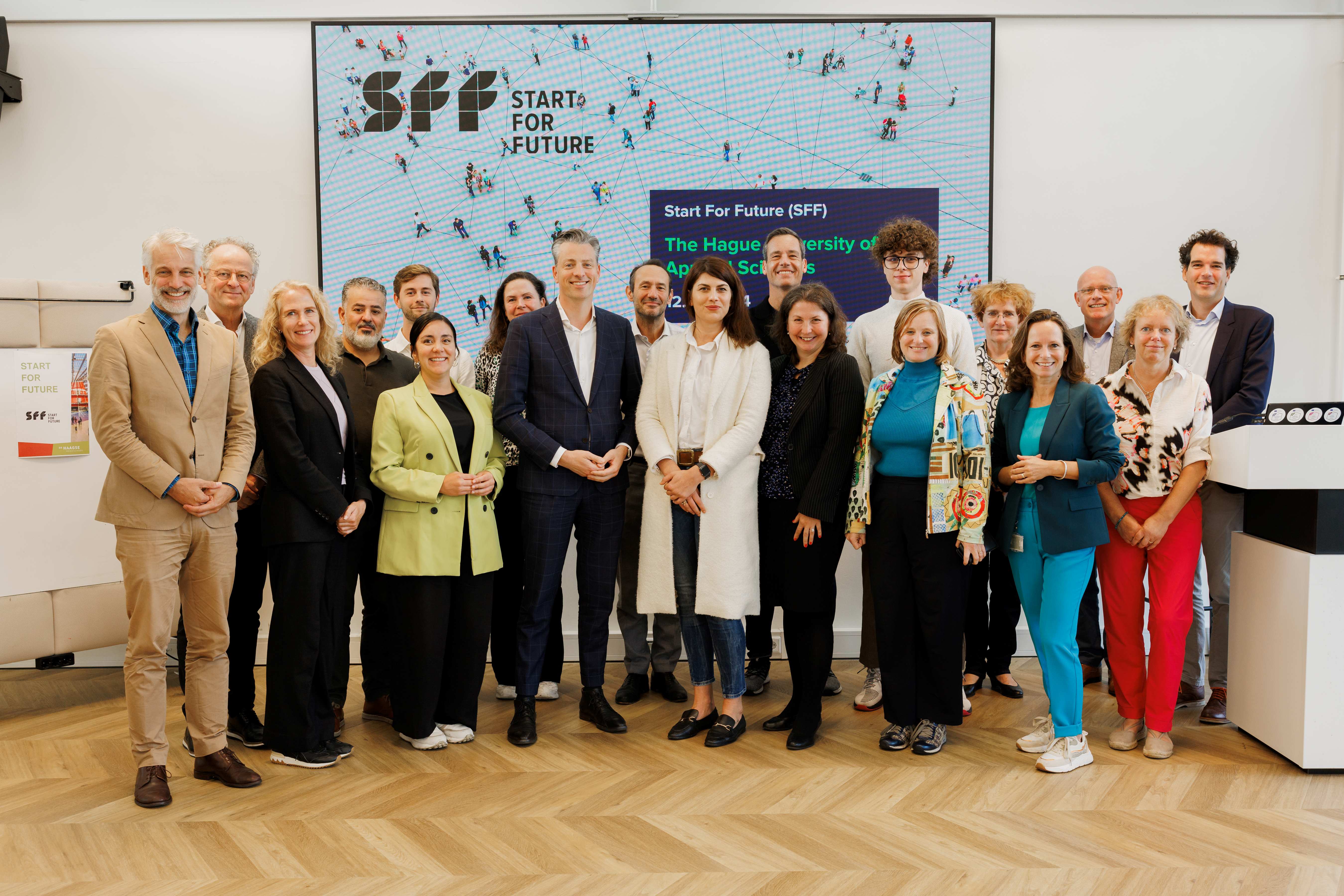 Groepsfoto van deelnemers aan het Start for Future (SFF)-evenement, poserend voor een groot scherm met het SFF-logo en informatie over The Hague University of Applied Sciences.
