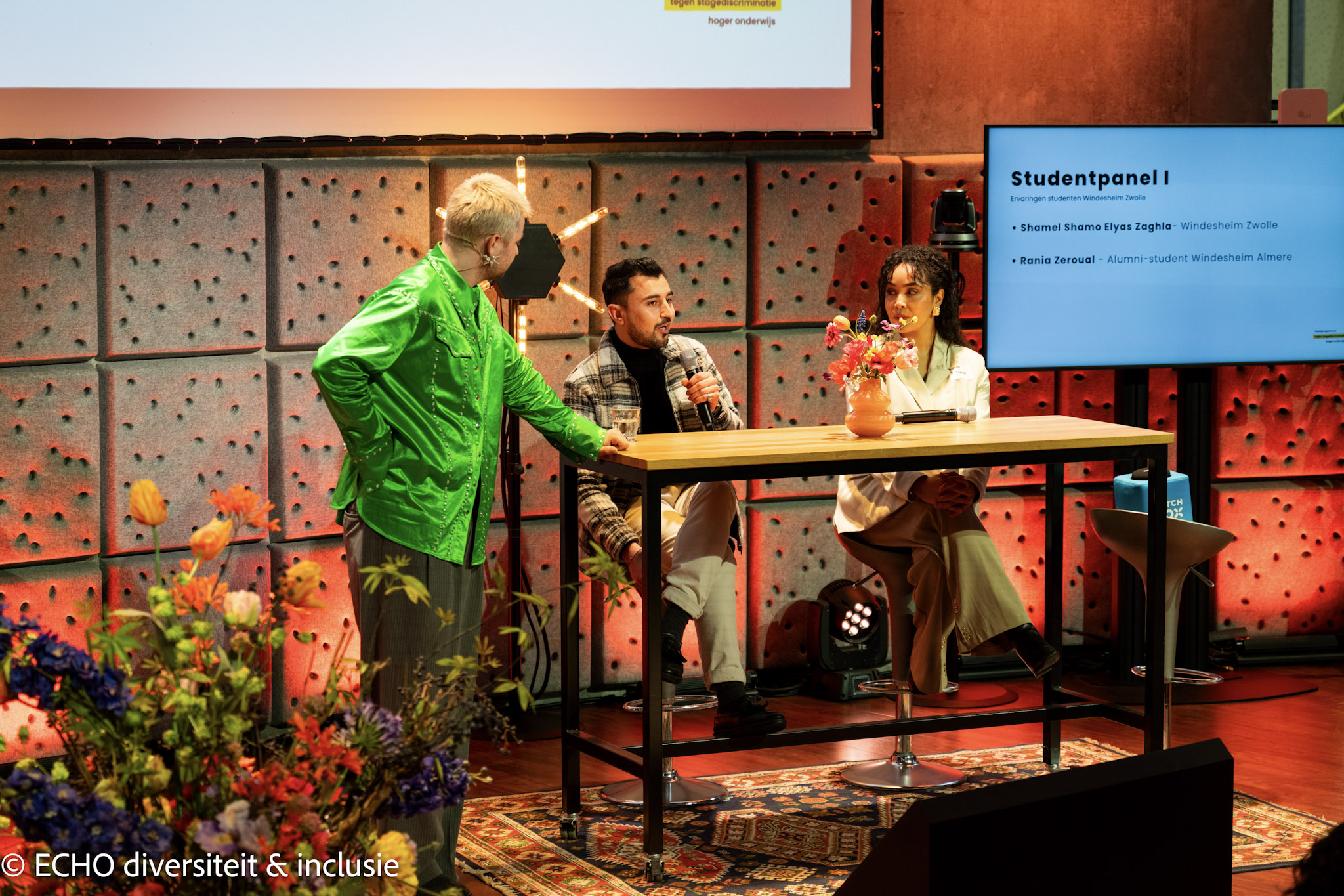 Paneldiscussie met drie deelnemers aan tafel tijdens een evenement over diversiteit en inclusie, met op de achtergrond een scherm waarop een studentenpanel wordt aangekondigd.