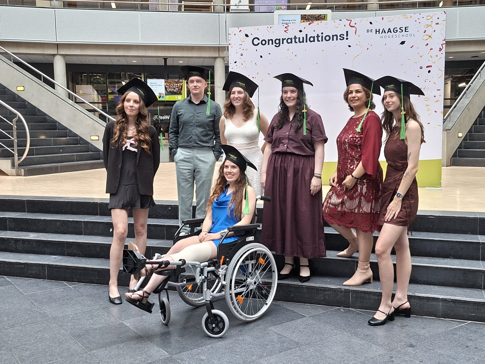 A group of graduates in graduation caps pose for a photo in the atrium of The Hague University of Applied Sciences, with one graduate seated in a wheelchair in the foreground.