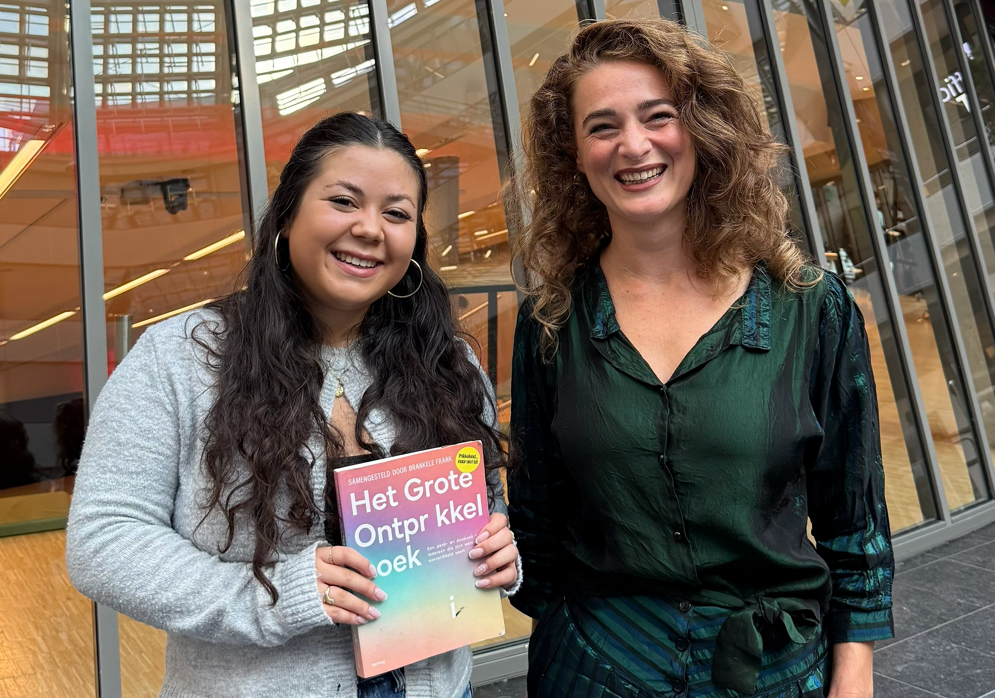 Twee personen staan naast elkaar in een gebouw met grote glazen ramen, waarbij een van hen het boek Het Grote Ontprikkelboek vasthoudt.